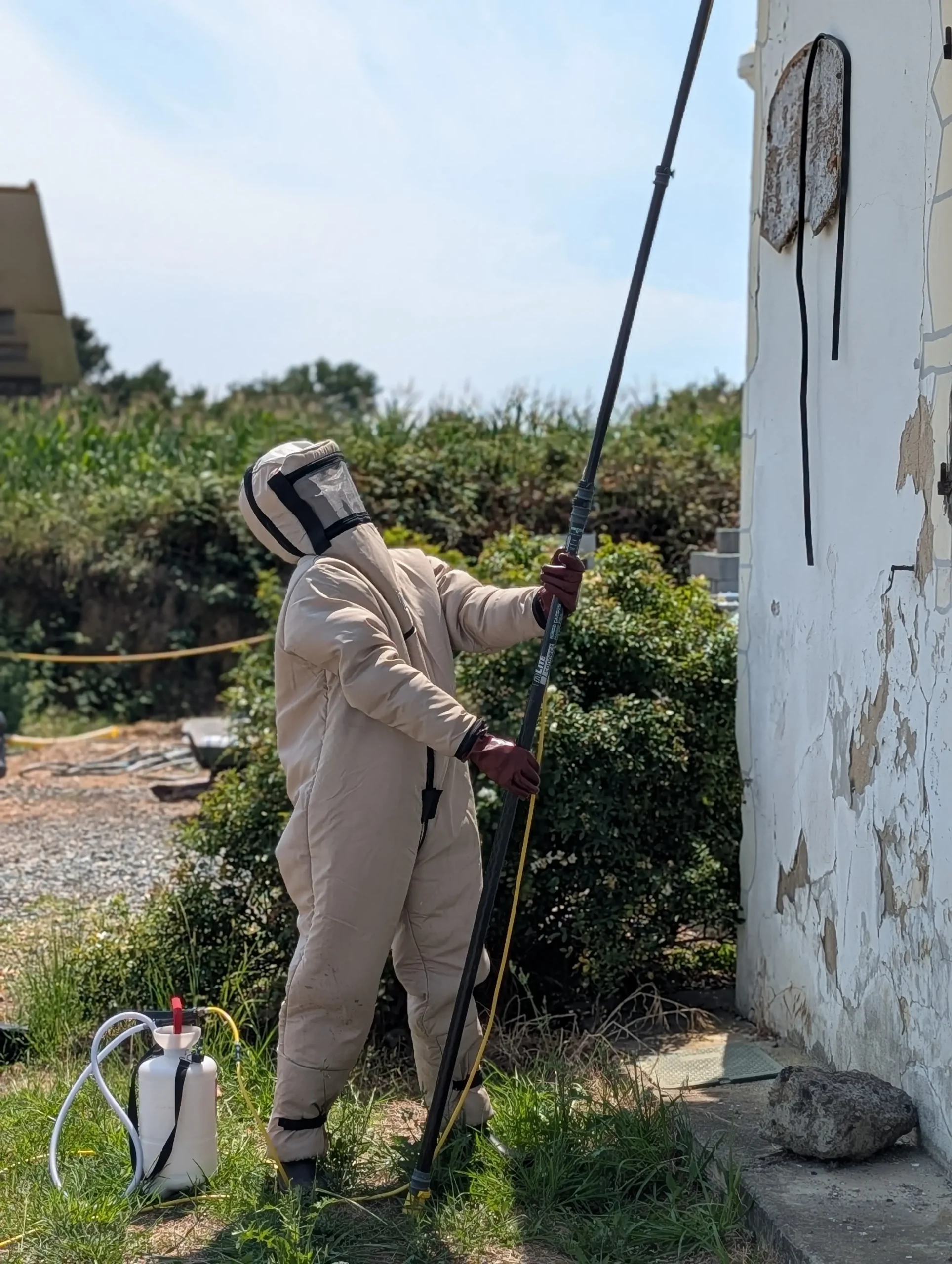 Person in beige protective suit with hood and gloves guiding a long spraying wand beside a white, peeling wall outdoors.